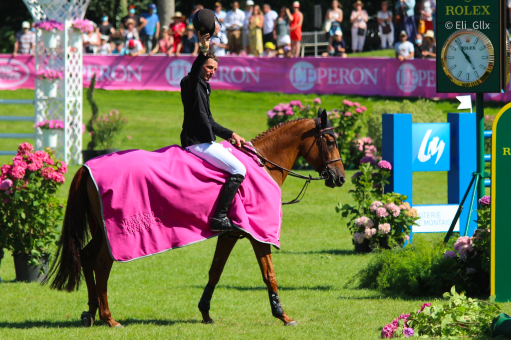 Kevin Staut waving. Competition on a brown horse with pink planket. The horse is walking slow. audience are looking. Rolex watch. Pink flowers.