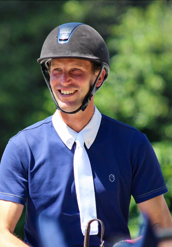 Kevin staut close up with a blue polo shirt and tie. Wearing a helmet and smiling. Green background. 